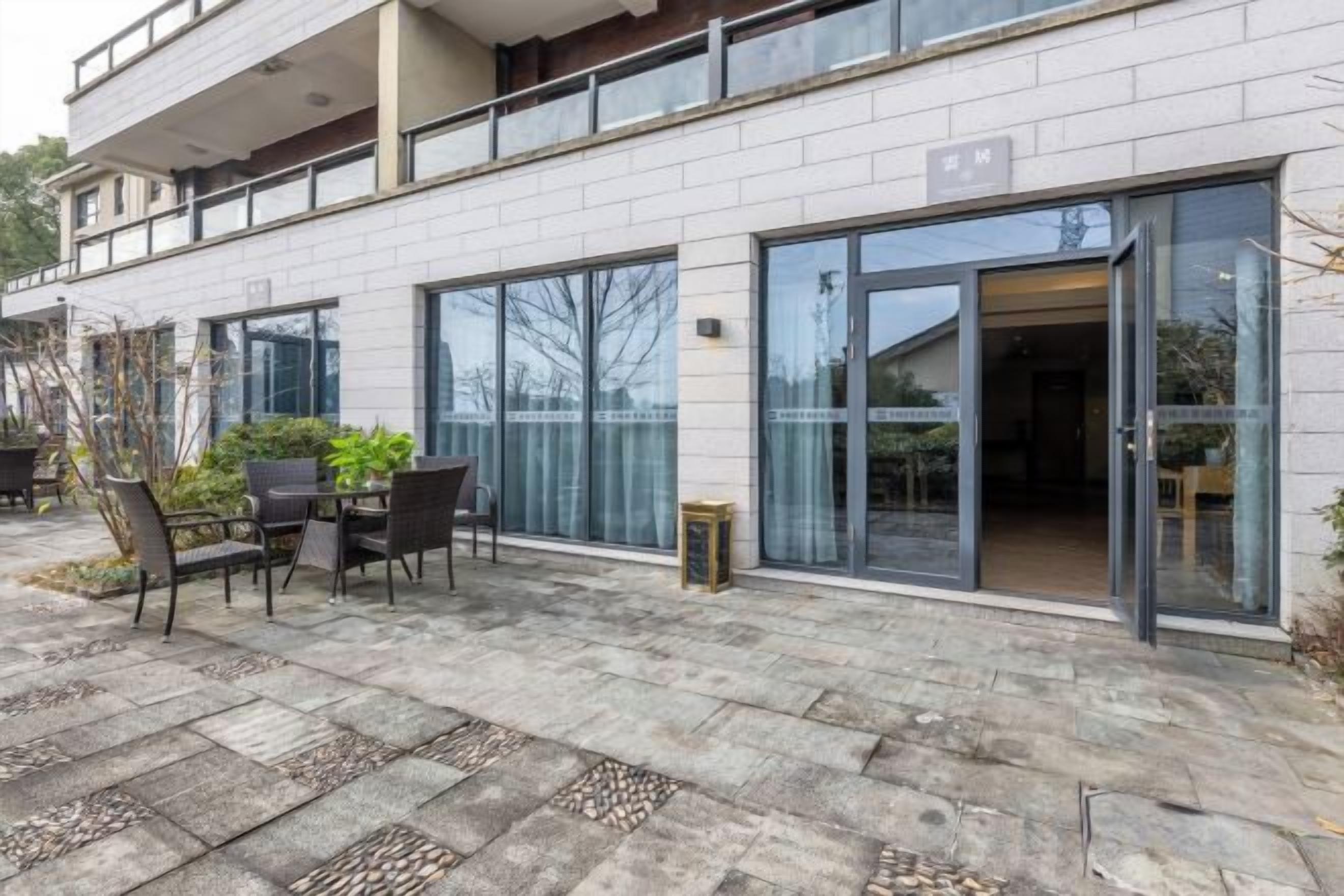 A modern building with a patio area featuring a table and chairs, large glass doors, and a stone-tiled floor.