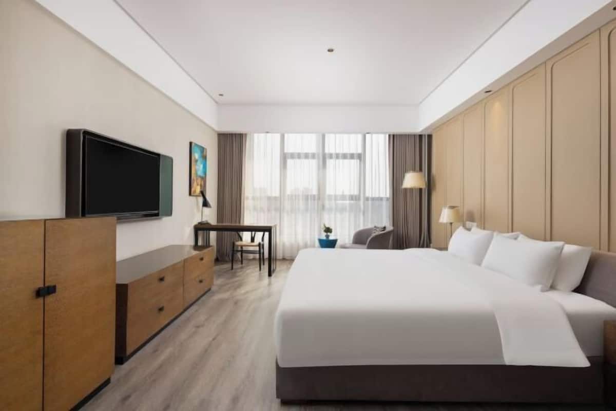Habitación de hotel con una cama grande, televisión, escritorio y vista de la ciudad a través de amplias ventanas.