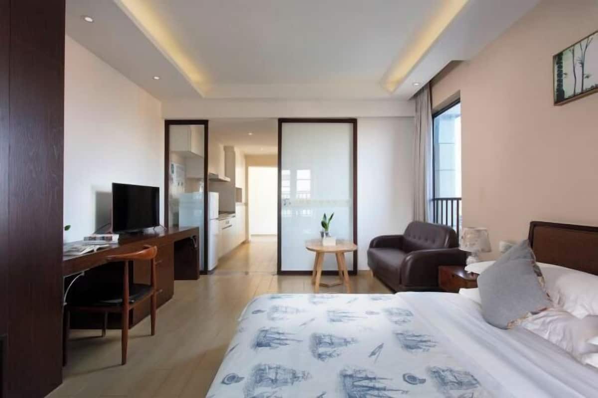 A hotel room with a bed, a desk with a television, a chair, a small table with a plant, and a balcony.