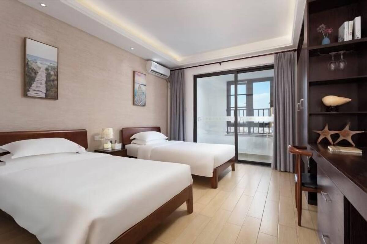 A hotel room with two beds, a large window, a balcony, and a wooden side table with decorative items.