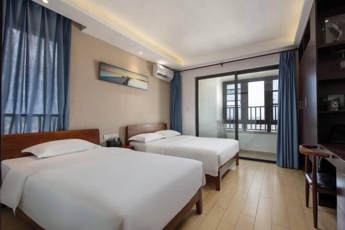 A hotel room with two beds, a desk, a chair, a large window with a view, and an air conditioning unit.