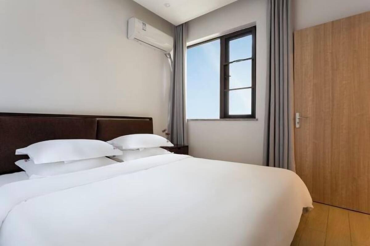 A hotel room with a bed, a window, an air conditioning unit, and a wooden wardrobe.