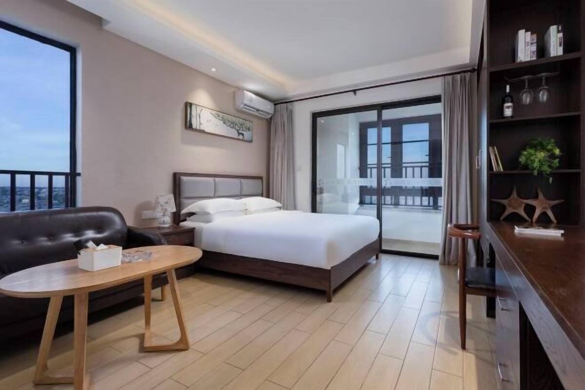 A hotel room with a bed, a wooden table, a sofa, a bookshelf, and a balcony with a view.