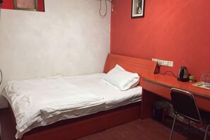 Un lit simple avec du linge de lit blanc, un mur rouge, un petit bureau avec une chaise et une poubelle.