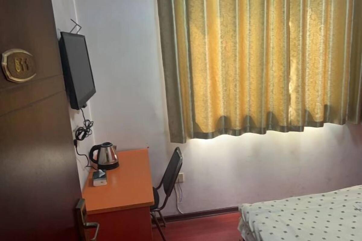 A room with a bed, a desk with a kettle, a chair, a television on the wall, and a window with curtains.