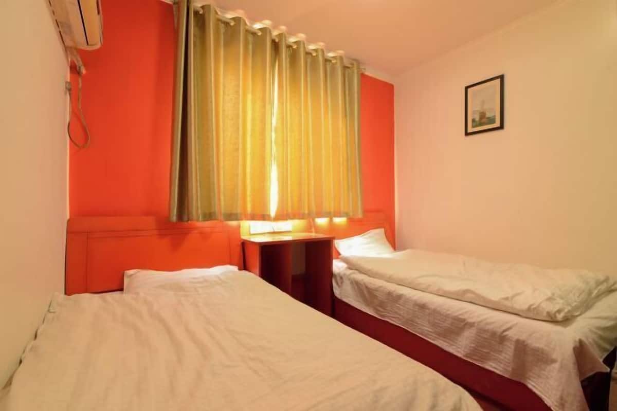 A room with two single beds, a small table, an air conditioning unit, and a window with curtains.