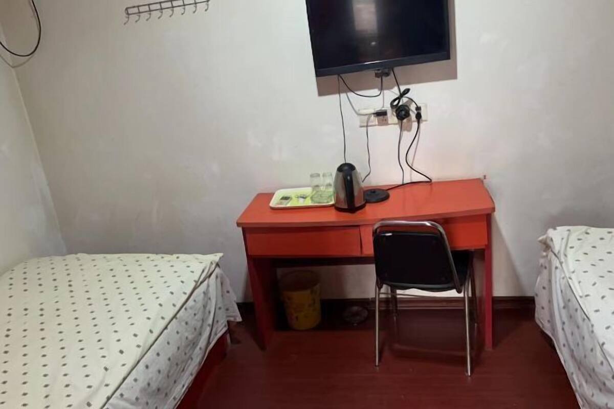 A small room with a single bed, a desk with a chair, a television, and a trash bin.