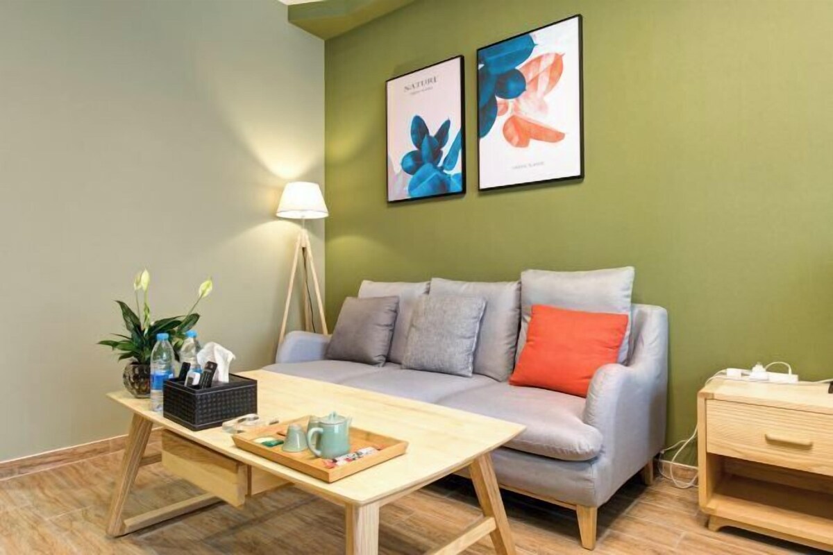 A room with a grey sofa, a wooden coffee table, a bedside table, a lamp, and two framed artworks on the wall.