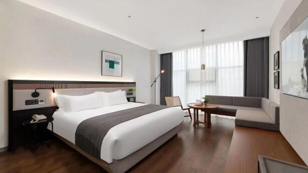 A modern hotel room with a large bed, a bedside table with a phone, a small round table, a sofa, and a large window with curtains. - Gujia Hotel (Xinxiang)