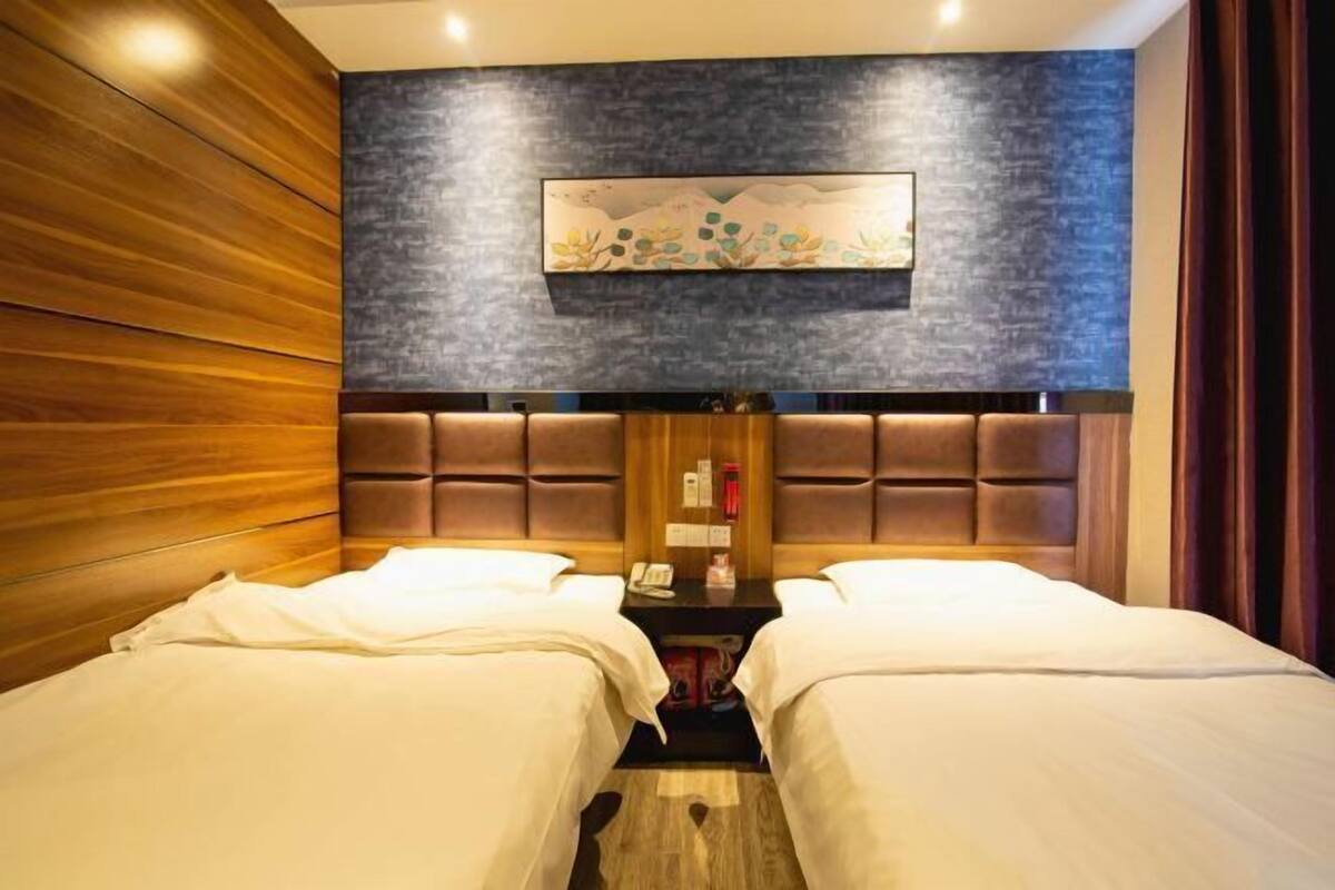 A hotel room with two beds, a wooden panel wall, a dark textured wall with a painting, and a brown headboard.
