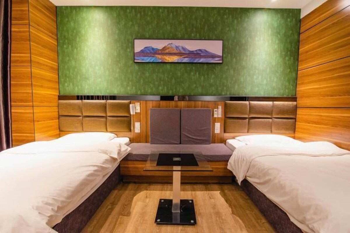 A hotel room with two beds, a central table, a green textured wall, and a painting above the bed.