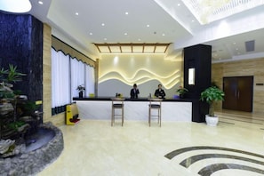 Un'area reception di un hotel dal design moderno, con una scrivania della reception, due sgabelli da bar, un bancone reception e un'ampia pianta in vaso, oltre a una parete decorativa.