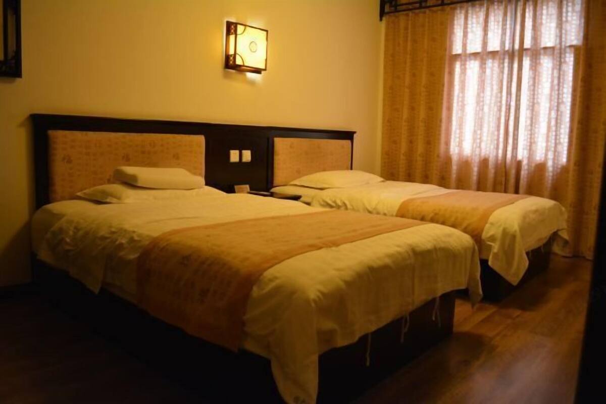 Une chambre avec deux lits, une table de chevet, une lampe fixée au mur et une fenêtre avec des rideaux.