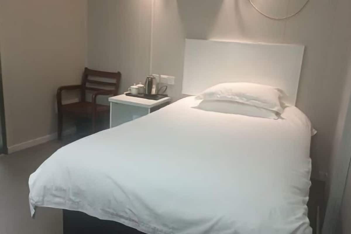A hotel room with a bed, a chair, a small table with a kettle, and a wall-mounted light fixture.