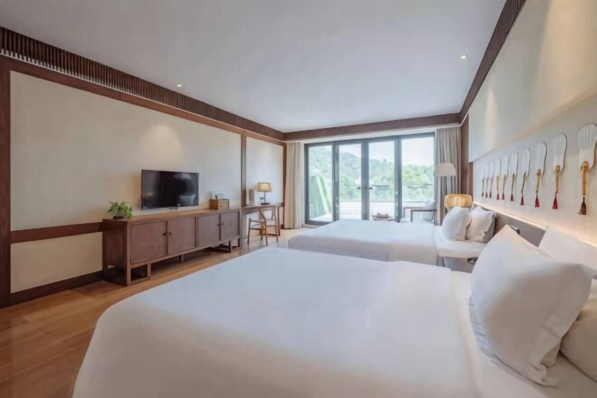 Habitación de hotel con dos camas, televisión, escritorio con lámpara y un ventanal grande.
