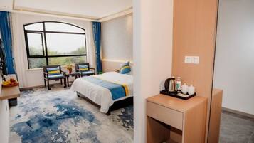 A hotel room with a bed, a desk with a mirror, a chair, a window with curtains, and a small table with toiletries.