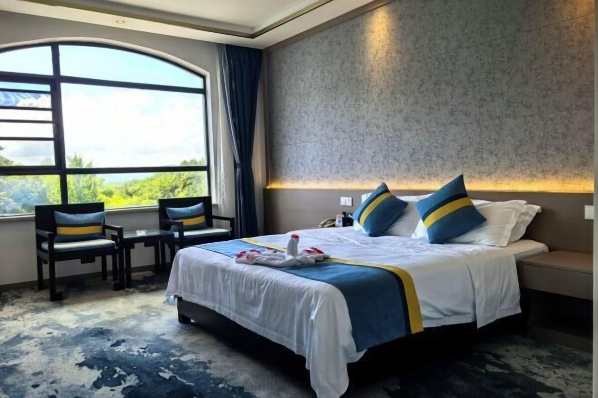 Habitación de hotel con una cama grande, dos sillones, una mesita y vistas a zonas verdes a través de un ventanal amplio.