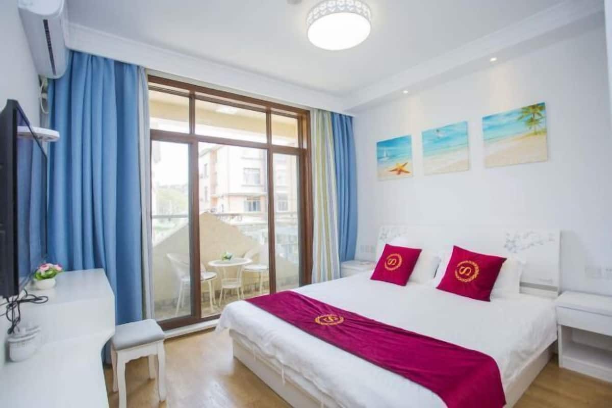 Une chambre d’hôtel avec un lit, un bureau et une chaise. Il y a trois tableaux au mur, une fenêtre avec des rideaux bleus et une vue sur l’extérieur.