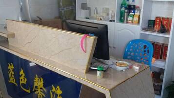 Mostrador de recepción con un monitor de computadora, teclado y ratón. Hay estantes con diversos objetos, incluyendo botellas y cajas.