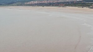 Egy széles strand, ahol a hullámok lágyan fodoroznak, a távolban egy város, és tiszta az ég.