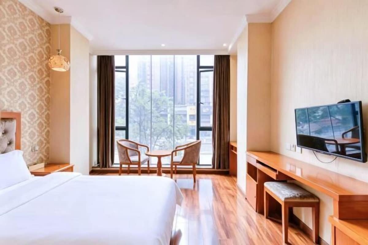 A hotel room with a large bed, a TV, a desk with a chair, and a view of the city through large windows.