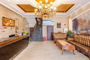 Um saguão de hotel com balcão de recepção, escada, lustre e uma pintura grande.