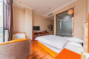Quarto de hotel com piso de madeira, cama com roupa branca, escrivaninha com televisão e uma janela grande.