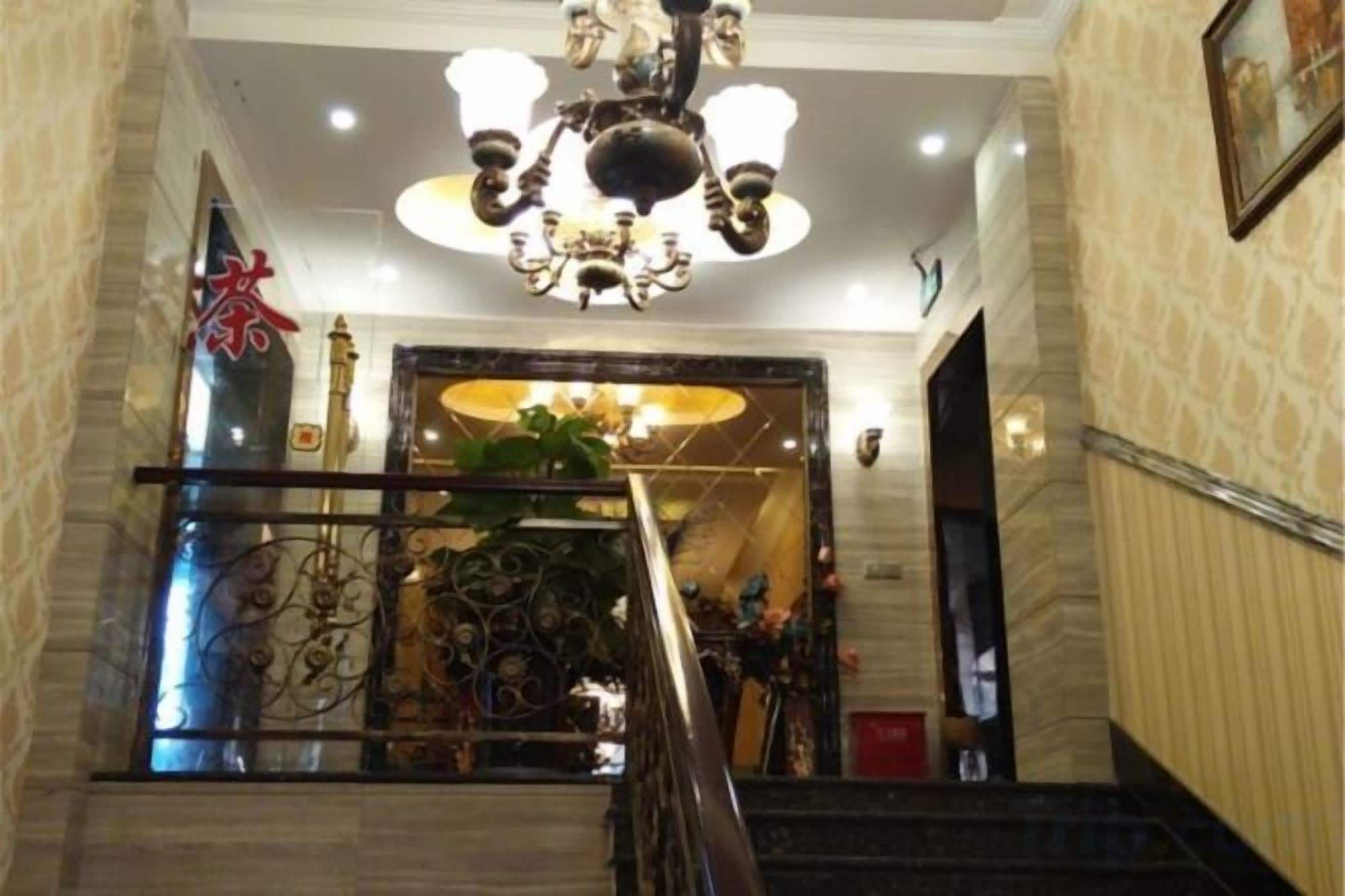 Una imponente escalera con candelabro, barandilla decorativa y vista a un salón con un espejo grande y una planta.