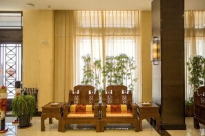 Een hotellobby met een houten zithoek, grote ramen met transparante gordijnen en een hoge pilaar met een lamp.
