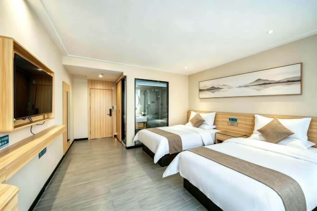 A hotel room with two beds, a television, a painting, and a bathroom visible through a glass door.