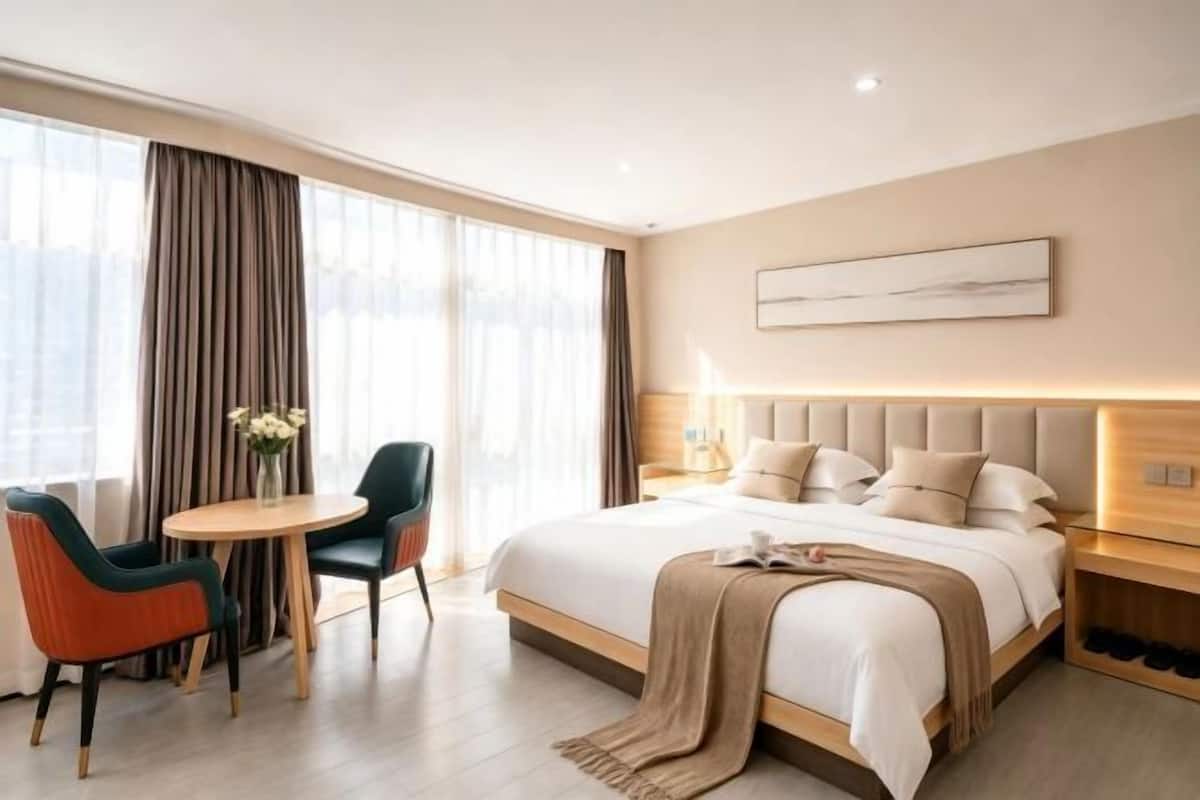 A modern hotel room with a large bed, a round table with chairs, and a view through large windows.