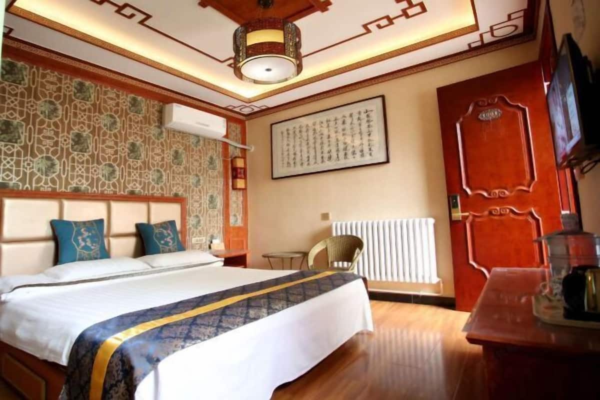 A hotel room with a bed, a wooden headboard, a wall with traditional patterns, a framed calligraphy piece, a radiator, and a television on a wooden stand.