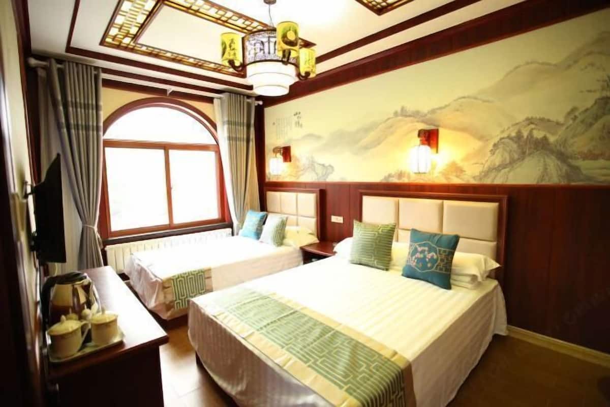 A hotel room with two beds, a large window with curtains, a traditional chandelier, and a mural on the wall.
