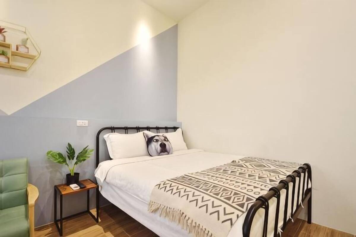 Un dormitorio con cama, mesita de noche, una silla, planta en maceta y estante en la pared.