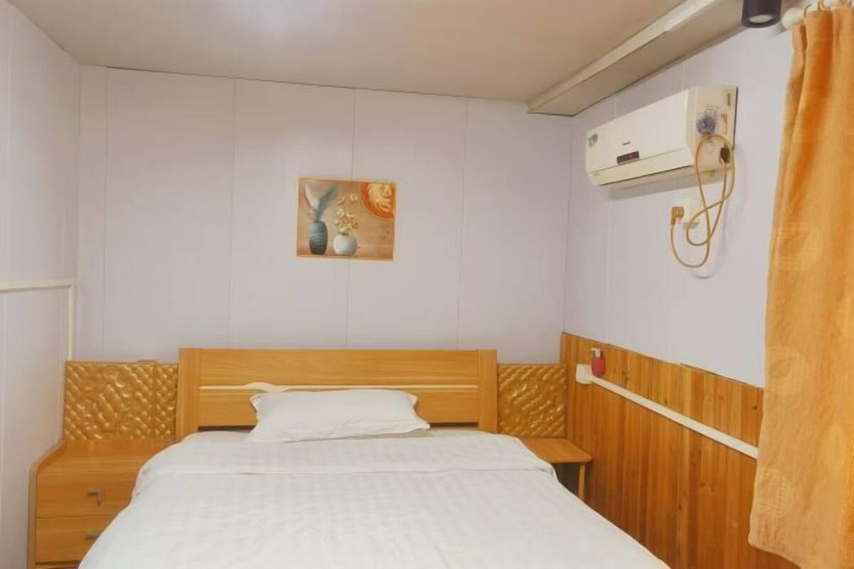 A bedroom with a wooden bed, a nightstand, a wall-mounted air conditioner, and a painting.