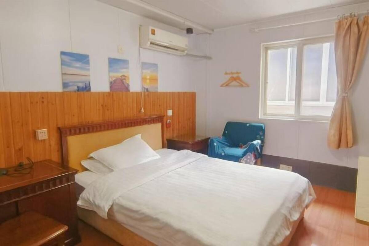 A hotel room with a wooden headboard, a bed with white bedding, a desk with a chair, a window with curtains, and wall art.