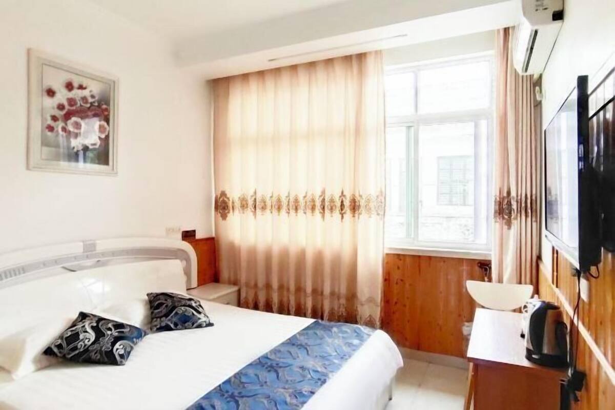 A hotel room with a bed, a bedside table with a kettle, a TV mounted on the wall, and a window with curtains.