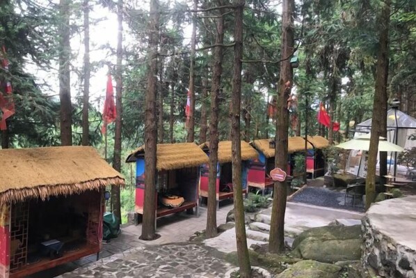 A forested area with thatched-roof huts, a stone pathway, and a flag. - Queen's Mountain Oxygen Heart Valley Mountain Resort (Lianshan)
