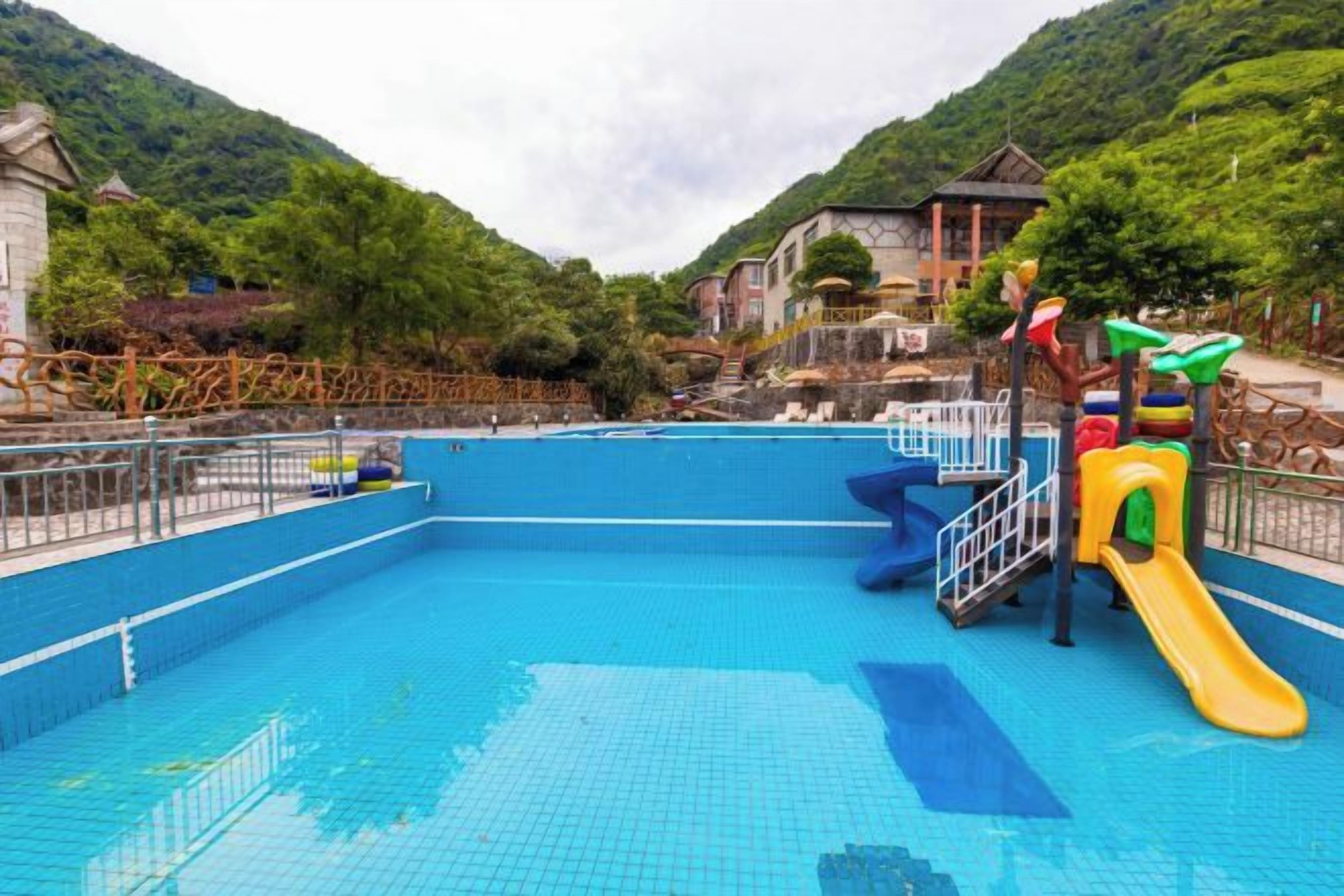 A swimming pool with a slide and a fenced area, surrounded by greenery and buildings.