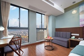 Hotellihuone, jossa on suuri ikkuna, työpöytä tuolilla, sohva, pieni pyöreä pöytä, jossa on hedelmiä, ja sänky.