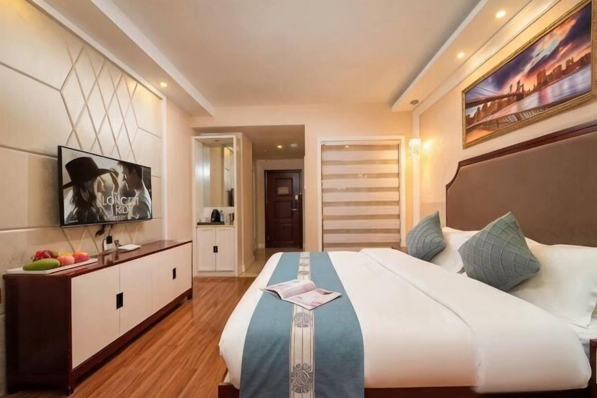 A hotel room with a large bed, a TV, a wooden dresser, and a cityscape painting.