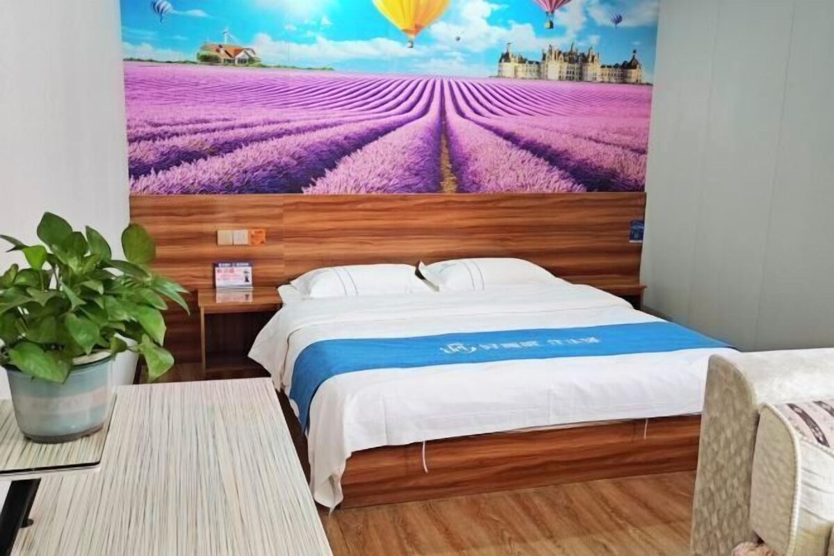 A hotel room with a bed, a wooden headboard, a nightstand, a bedside table, a potted plant, and a wall mural of a lavender field with hot air balloons.