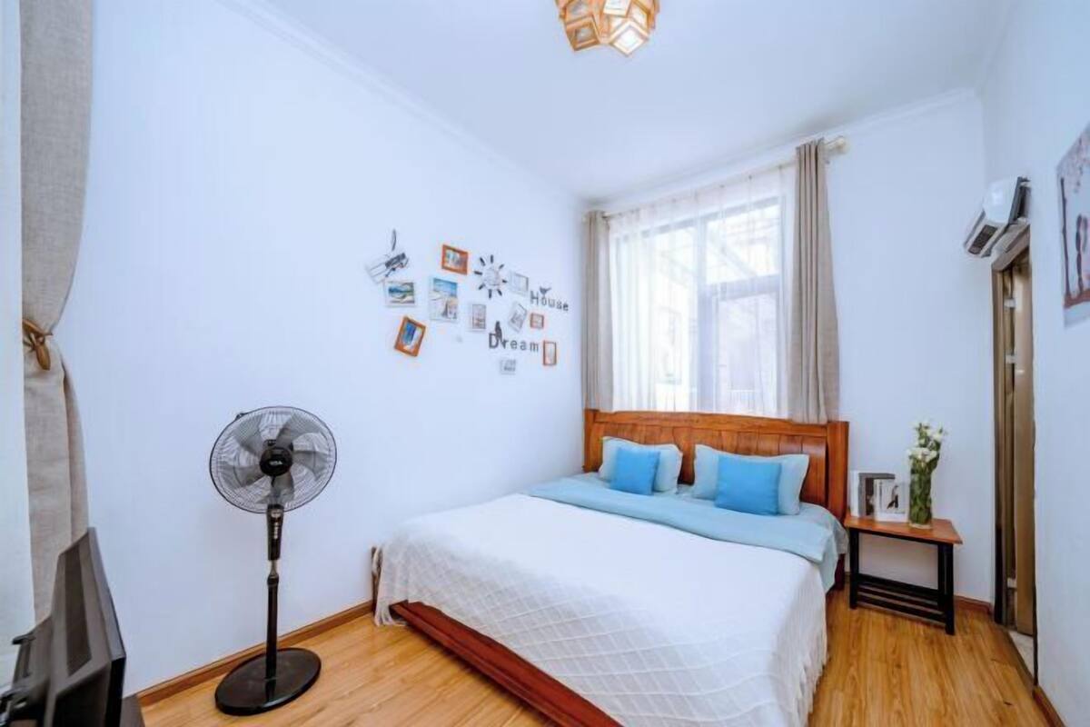 A bedroom with a bed, a standing fan, a bedside table with a lamp, and a window with curtains.