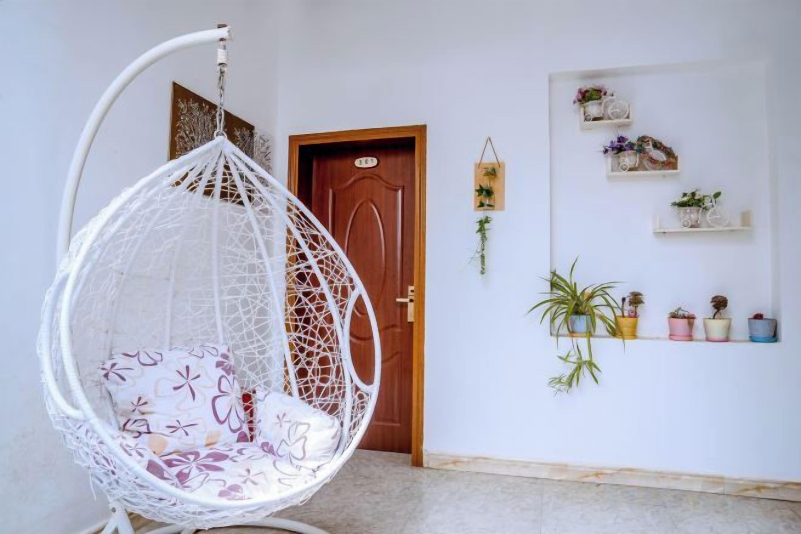 A white hanging chair with a cushion, a wooden door, and a wall with shelves holding potted plants and decorative items.