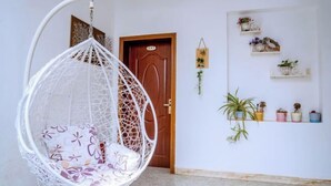 Une chaise suspendue blanche avec un coussin à motifs floraux, dans une pièce dotée d’une porte en bois, d’une étagère murale avec des plantes en pot et d’une petite plante suspendue.