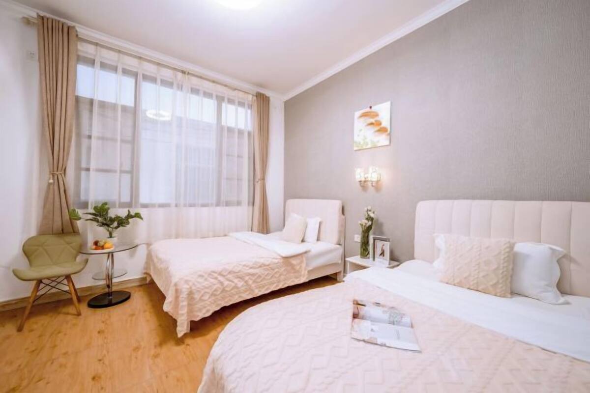 A hotel room with two beds, a small table with a plant, a chair, and a window with curtains.