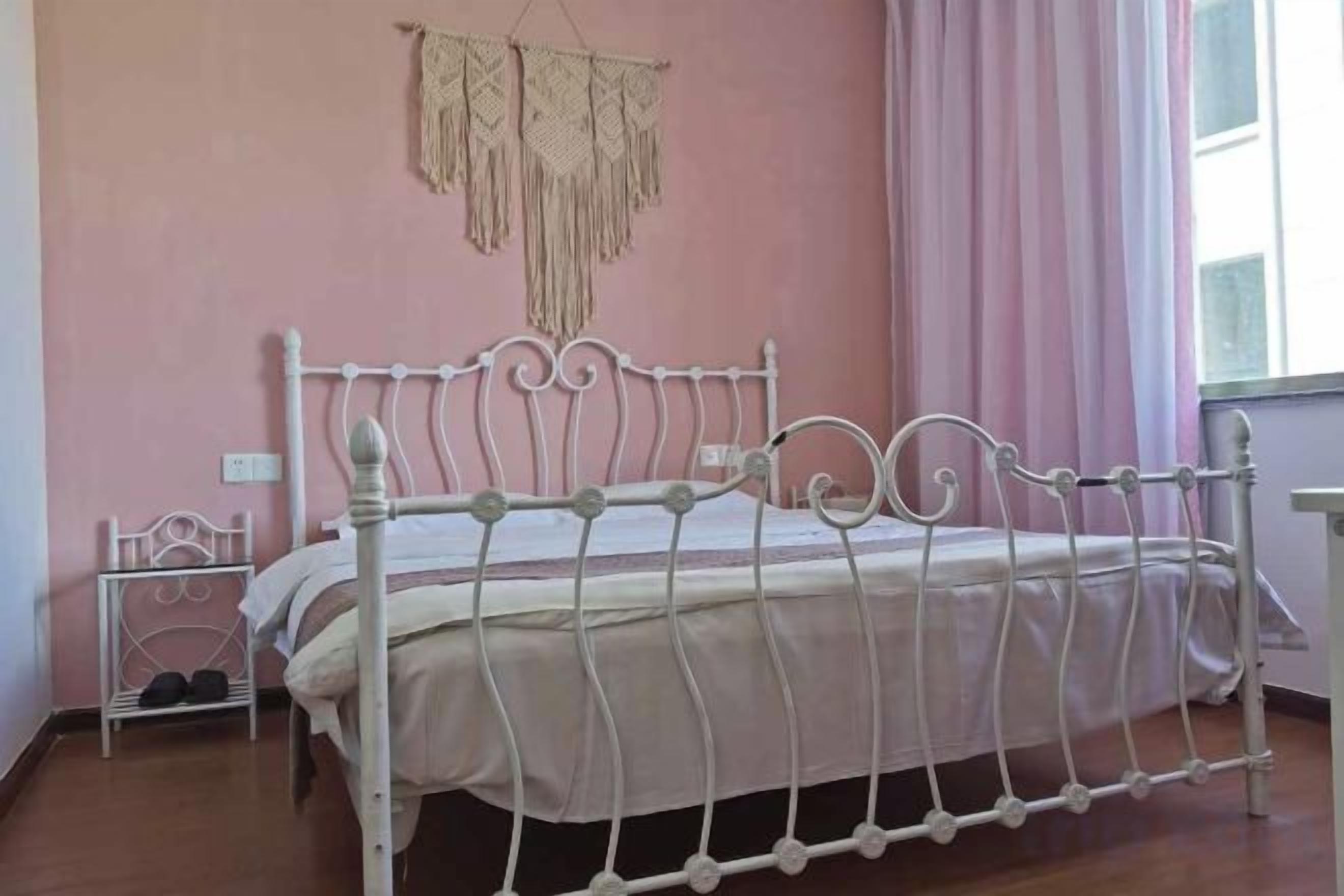 A white metal bed in a room with pink walls, a small bedside table, and a window with curtains.