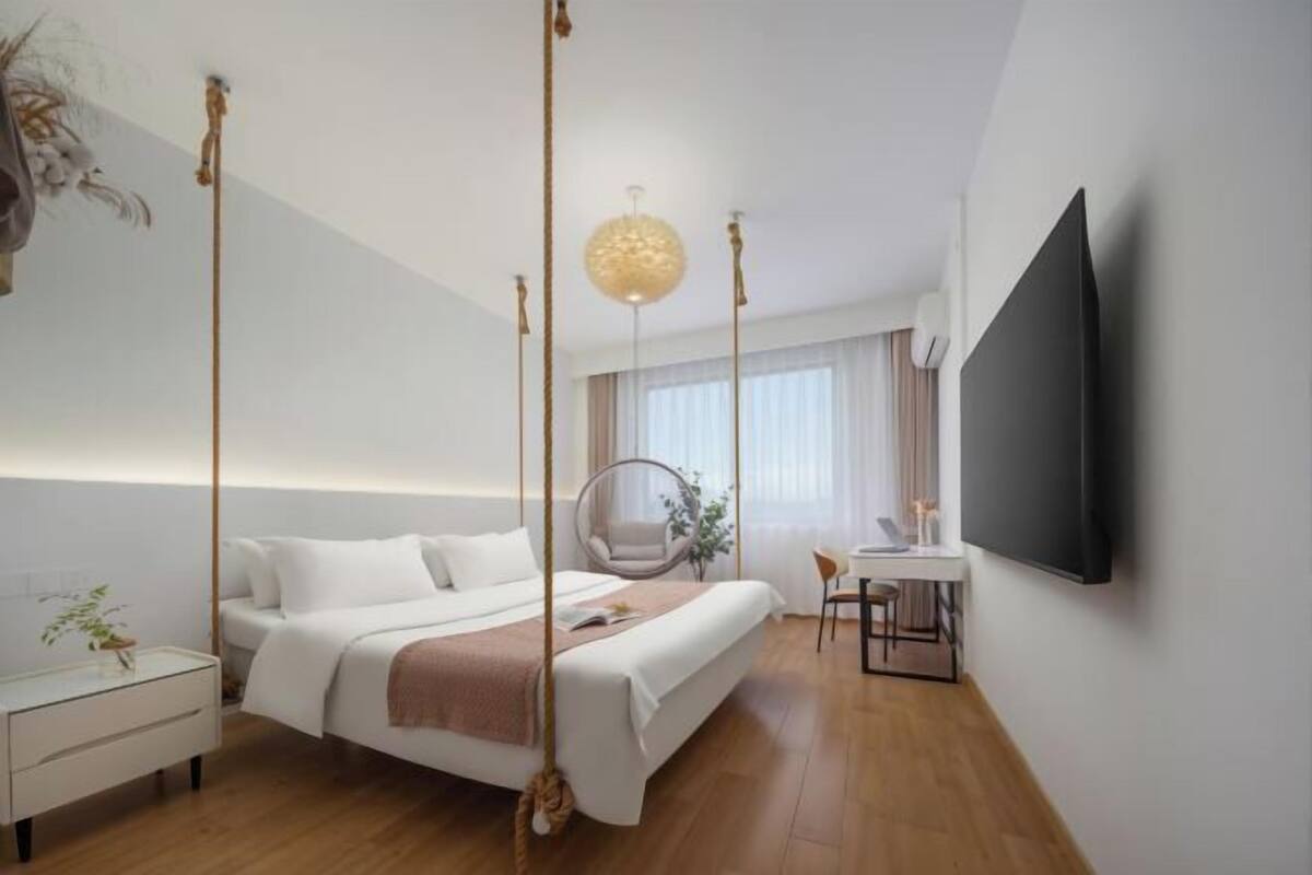 A modern hotel room with a bed, bedside table, hanging chair, desk, and a TV mounted on the wall.