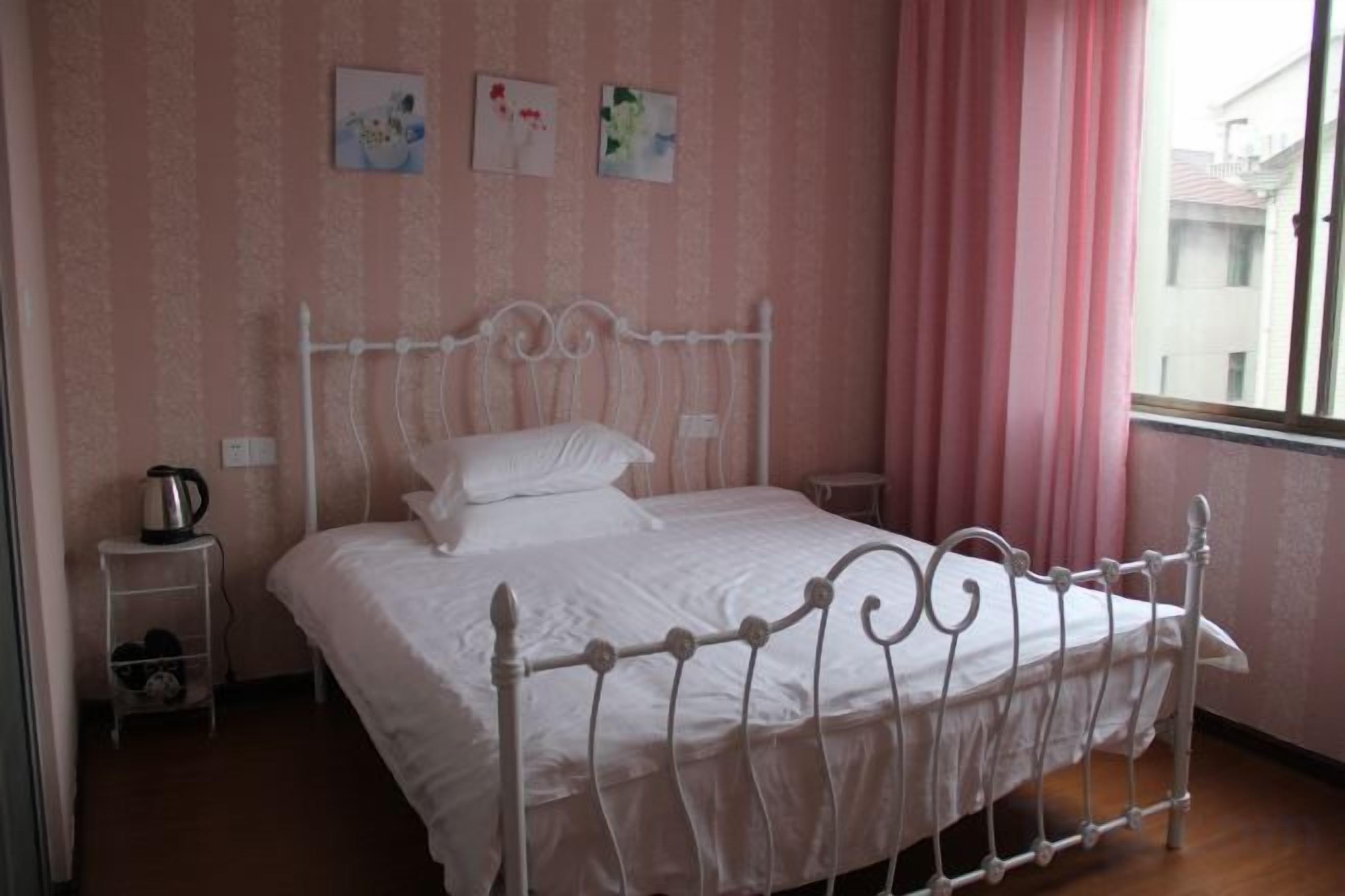 Un llit ben fet amb roba de llit blanca en una habitació amb paper pintat rosa i una finestra amb cortines.