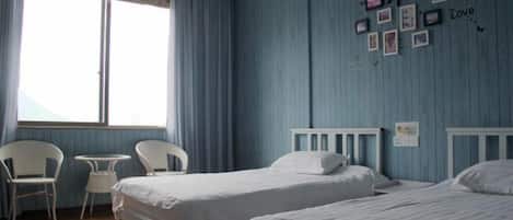 A room with two beds, a small table, and chairs. There is a window with curtains, and a wall with framed pictures.